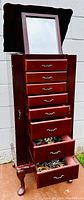 Full-height view of red-brown wooden jewelry armoire with mirror and drawers.