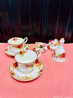 Two Royal Albert Old Country Roses tea cups with matching saucers, pair of floral candlestick holders, and a bell arranged on a pink surface against a dark backdrop, showing the set as a whole.