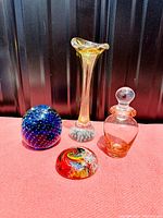 Four vintage studio art glass pieces including a blue bubble paperweight, tall vase, amber perfume bottle, and colorful dome paperweight on red cloth against dark background.