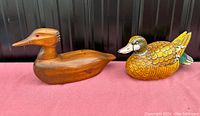 Two wooden carved duck decoys displayed on a red cloth with a dark background, showing a side view of both decoys.