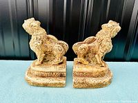 Pair of vintage hard plaster foo dog bookends shown facing each other on tiered bases against a black background.
