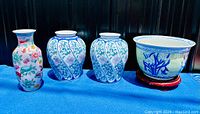 Overview of four Oriental ceramic items: two bulbous vases, one tall slender vase with colorful flowers and birds, and one planter on a wooden base arranged on a blue cloth.