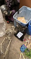 Photo of two plastic skeletons on floor next to storage bin and witch figure head with crystal ball.