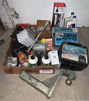 Overview of assorted automotive parts including oils, filters, snow chains, seat belts, and tow hitch parts arranged in a box and on the floor.
