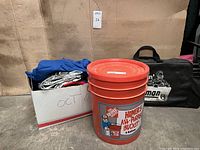 Two tent footprints in blue and silver, red mylar tarp, orange nesting bowls inside a large labeled cardboard box and a large orange bucket labeled Home Depot 5 gallon bucket.