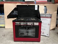 Front view of red Camp Chef oven with lid open, showing two burners and oven door