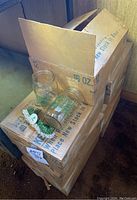 Opened box showing seven unopened boxes stacked. One box is open showing some canning jars with green Evergreen School District logos and green and white garters.