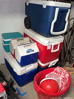 Seven coolers stacked; includes wheeled Coleman cooler, two Little Playmate small coolers, other various sized coolers, and a red bucket with picnic items