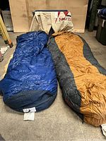 Two down sleeping bags laid out on floor; Marmot in burnt orange and black, Ascente in blue and black.