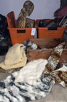 Large skeleton seated in orange storage bin behind assorted fabrics and a wooden box, showing full scene with Halloween decor items.