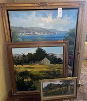 Three framed paintings stacked against a wall showing coastal and barn scenes with one small, one medium, and one large painting.