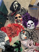 Assorted Halloween masks with various scary designs laid on floor including a Scream mask, zombie-style mask, purple hair skull, red monster face, and grotesque bearded face.