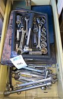 Drawer with combination wrenches, a ratchet with black handle, and assorted sockets lying loosely and in a black plastic tray.