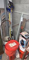 Photo shows collection of items including long metal poles, Simer sump pump in box, Bosch digital level, aluminum T-square, orange buckets, and rope all resting against a concrete block wall.