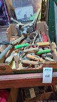 Box of assorted hand tools including hatchets, curved saw, hay hooks, and more in a cardboard box
