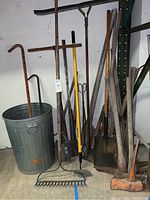 Photo showing assorted yard tools including metal rake, shovels, pick axes, crow bars, hoe, large pruners, metal rulers; handles appear in varying condition; one aluminum garbage can visible.