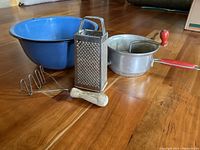 Photo showing all four antique kitchen items including blue metal bowl, metal box grater, wooden handle metal masher, and red handle potato ricer on wood floor.