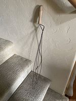 Photo of antique rug beater showing full length from wooden handle to wire loops resting on carpeted stairs, illustrating size and condition.