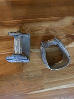 Photo showing two antique wooden stirrups placed on a wooden floor, one rectangular with metal frame and wooden foot block, the other rounded with wooden sides and metal fittings, both weathered and used.