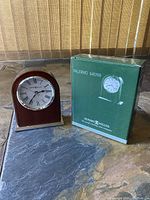 Front view of the Howard Miller Palermo clock next to the original green box showing the clock face with Roman numerals and brass base.