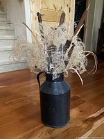 Front view of dried arrangement showing cattails, grasses, and feathers in black metal pitcher on wooden floor near stairs.
