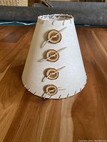 Front view of handmade lampshade showing cream paper finish, leather lacing along edges, and four wooden buttons with leather cord detail.