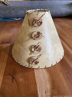Front side view of the handmade lampshade showing beige/tan paper finish, four wooden buttons on the side with leather lacing through them, and additional leather lacing along the bottom edge.