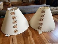 Two matching large handmade lampshades with cream paper finish, leather lacing, and wooden buttons, placed side by side on a hardwood floor showing front and side view.