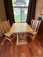 Log style dining table with solid pine top and two log dining chairs around it, placed on wood floor near window with brown curtains.