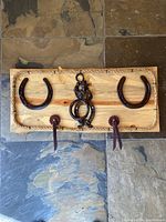Top view of the rustic towel rack showing pine wood backing, rope border, two horseshoes mounted on either side, central cowboy ring decoration, and two nails with leather strap hooks at the bottom.