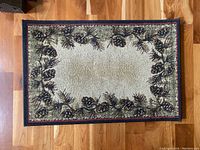 Top-down photo of rectangular indoor area rug with detailed pinecone and pine needle border on beige background, flooring visible underneath.