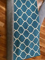 Rolled partly unrolled teal blue area rug with white interlocking geometric lines visible, shows wear and indentations from furniture.