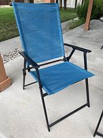 Front angled view of a teal blue outdoor folding chair with black metal frame and armrests on a concrete patio.