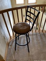 Photo of black metal bar stool with ladder back, beige round sueded cushioned seat. Positioned near wooden railing on landing with natural light from windows.