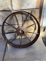 Front view showing the full circular shape, metal spokes, and textured weathered surface of the rustic wheel leaning against a stone wall.