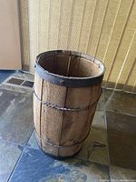 Small antique wooden barrel standing upright with open top, showing wooden staves and metal rings. The bottom metal ring is visibly dented.