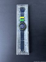 Swatch Scuba 200 diver watch packaged in original clear plastic case with water bubble graphics. Dark blue watch band with two rubber loops in green and yellow, "Swiss Made" and water resistance details visible.