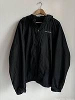 Front view of black men's Columbia jacket hanging on wooden hanger showing full jacket with hood and zippered front.