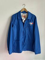 Front view of a bright blue Levi's jacket with Toronto Blue Jays logo on left chest pocket, button snap closure, hanging on wooden hanger against white wall.
