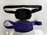 Top view of two waist packs side by side, black one on top and purple one below on white background.