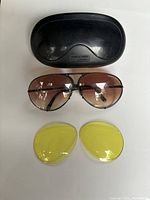 Black hard sunglasses case with embossed Porsche Design logo and Made in Austria mark, displayed with brown gradient lenses attached to sunglasses and separate yellow tinted lenses.