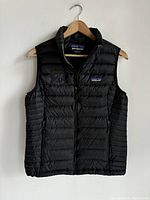 Front view of the black Patagonia down vest hanging on a wooden hanger. Visible crest monogram embroidered on the left side and Patagonia logo patch on the chest.