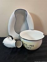 Photo showing all three vintage white enamel bathroom items: bed pan, pee pee pot labeled clearly, and urinal with handle, displayed against a wall.
