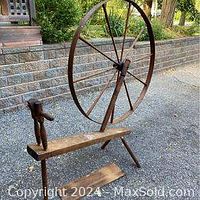 Large antique solid wood spinning wheel outdoors on pavement near stone retaining wall and garden. Wheel is intact; base is disassembled.