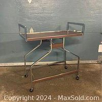 Front view of vintage metal tea cart with wheels, showing top rectangular tray with raised edges and tubular frame with angular supports on caster wheels.