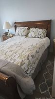 Front side view of queen size wooden bed with floral bedding and pillows, bedside table with lamp visible.