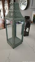 Tall green metal candle holder with intricate cutout designs and glass panels shown from an angle with smaller dark candle holder visible in background