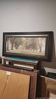 Framed scenic picture depicting a tree-lined pathway, in a dark frame with light inner border, hung on a wall.