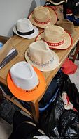 Top view of six straw fedora hats and two white fedora hats on a wooden table showing colorful decorative bands and embroidered text.