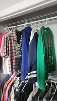 Full view of seven men's shirts and sweaters hanging on a clothing rod including a red and white plaid shirt, black patterned sweater, and a green Christmas sweater.